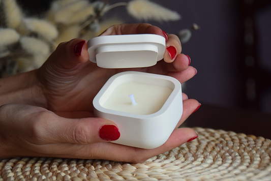 Vela decorativa en caja pequeña con tapa sujetada por manos de modelo en fondo rustico.