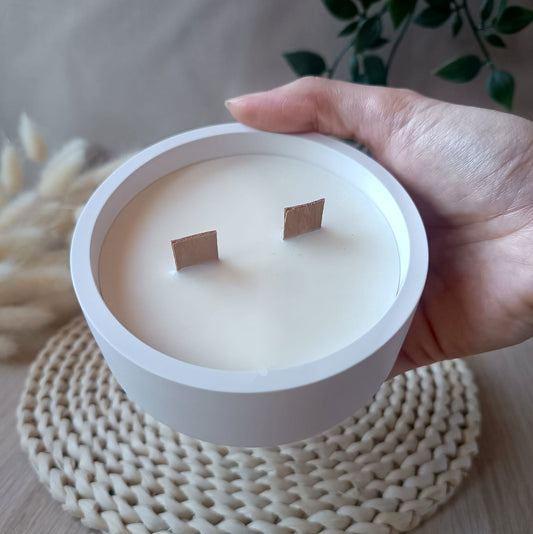 Vela decorativa grande de soja en recipiente blanco liso con doble mecha de madera sostenida en la mano y fondo neutro