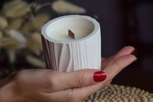 Vela decorativa en recipiente con formas organicas con mecha de madera, sujeta por mano de modelo y fondo rustico.