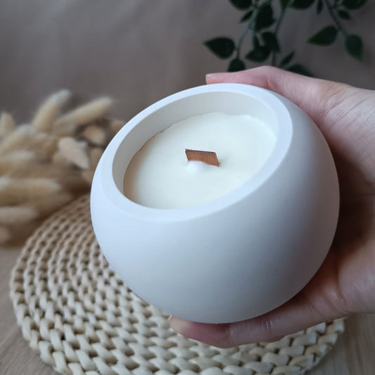 Vela grande de soja en recipiente esférico blanco liso de resina mineral con mecha de madera sostenida en la mano y fondo neutro