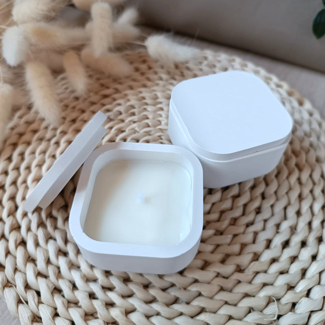 Dos velas decorativas de soja en recipiente cuadrado blanco con tapa y mecha de algodón sobre soporte de fibras naturales y fondo neutro