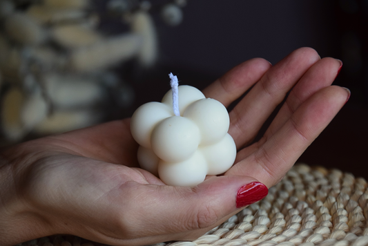 Vela quadrada con burbujas y pabilo de algodon, sujetada por mano de modelo con fondo rustico. 