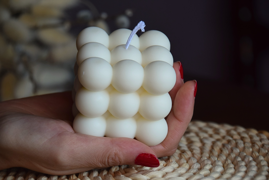 Vela decorativa quadrada con burbujas y pabilo de algodon, sujetada por mano de modelo en fondo rustico.
