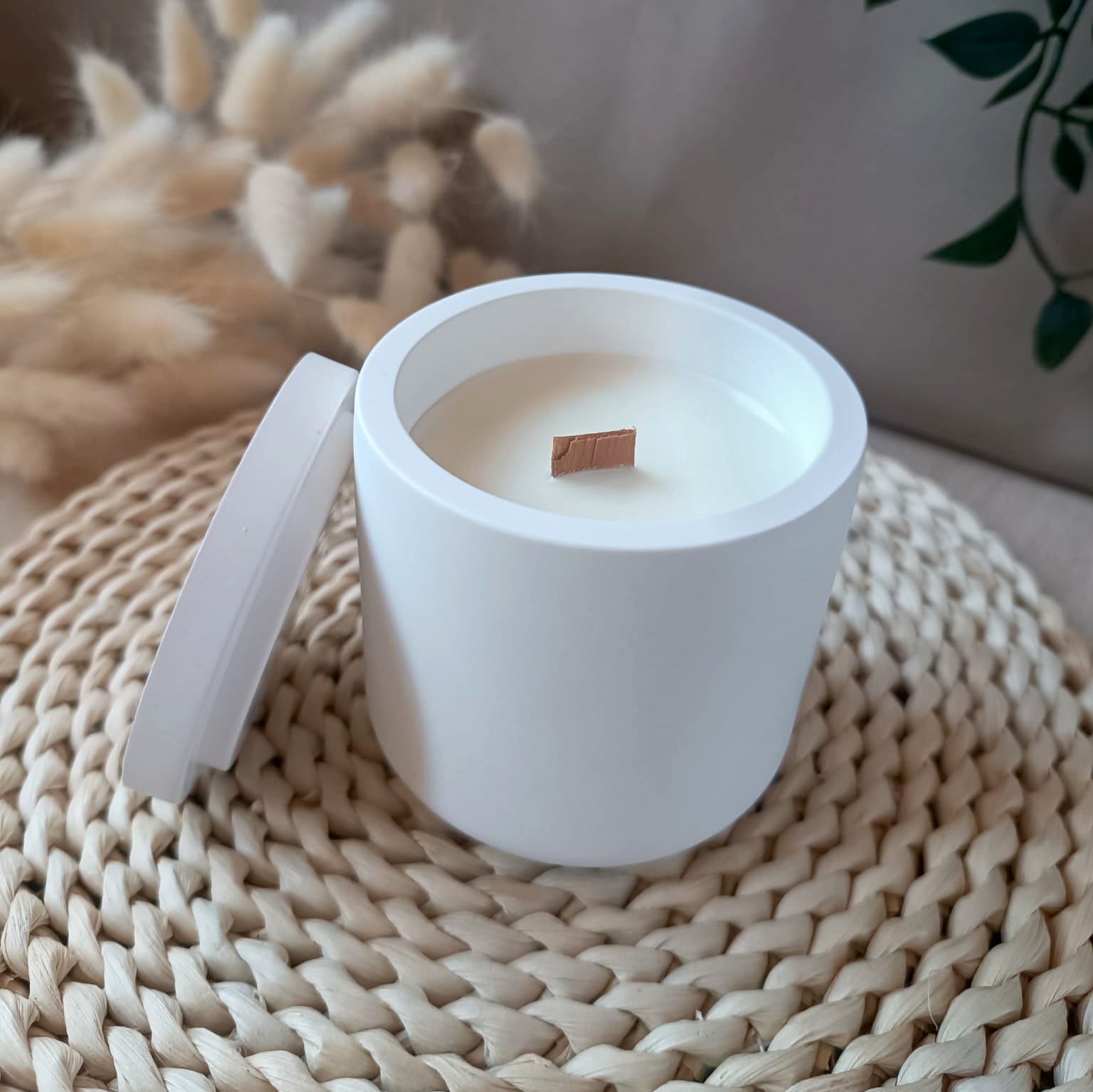 Vela decorativa de soja en recipiente blanco liso de resina mineral con mecha de madera y tapa sobre base de fibras naturales y fondo neutro