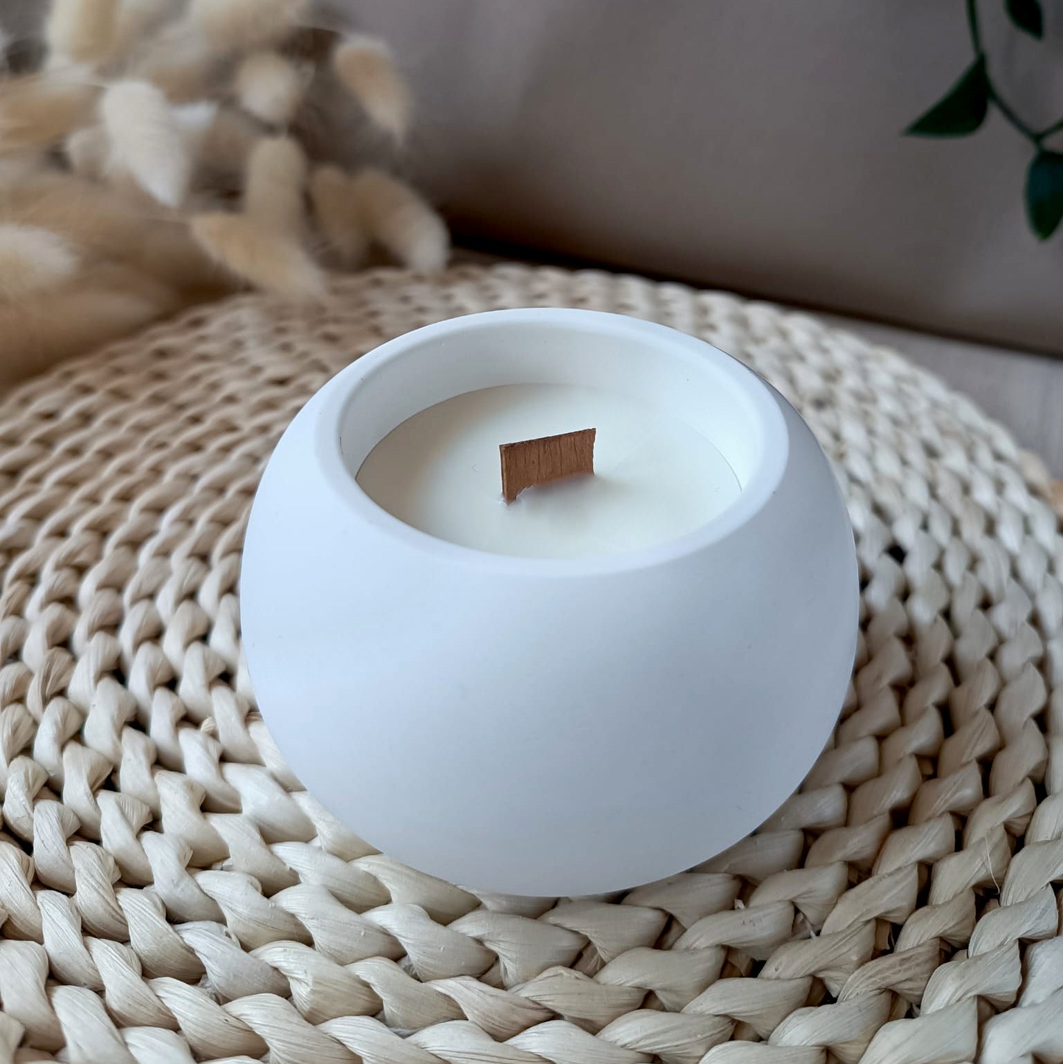 Vela decorativa mediana redonda de soja en recipiente blanco liso de resina mineral con mecha de madera sobre base de fibra natural y fondo neutro