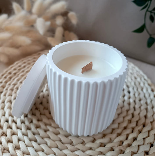 Vela decorativa de soja en recipiente blanco de resina mineral con textura de rayas verticales, mecha de madera y tapa a juego sobre base de fibras naturales y fondo neutro