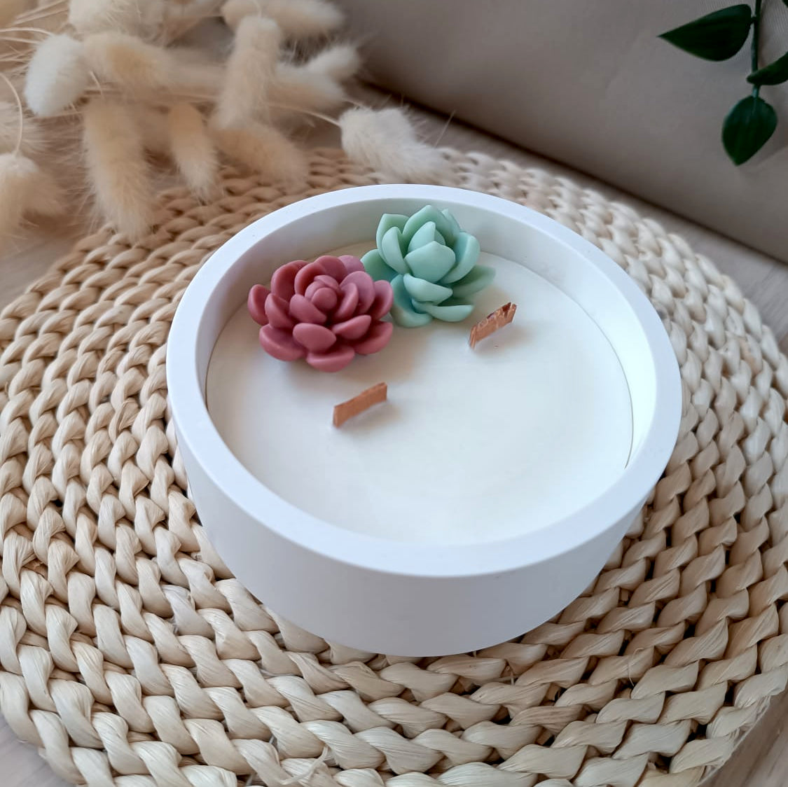 Vela decorativa de soja en recipiente de resina mineral de color blanco con suculentas y doble mecha de madera estilo natural sobre base de fibras naturales y fondo neutro