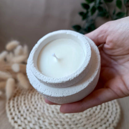 Vela decorativa de soja en recipiente blanco con textura rugosa efecto lava y mecha de algodón sostenida en la mano con fondo neutro