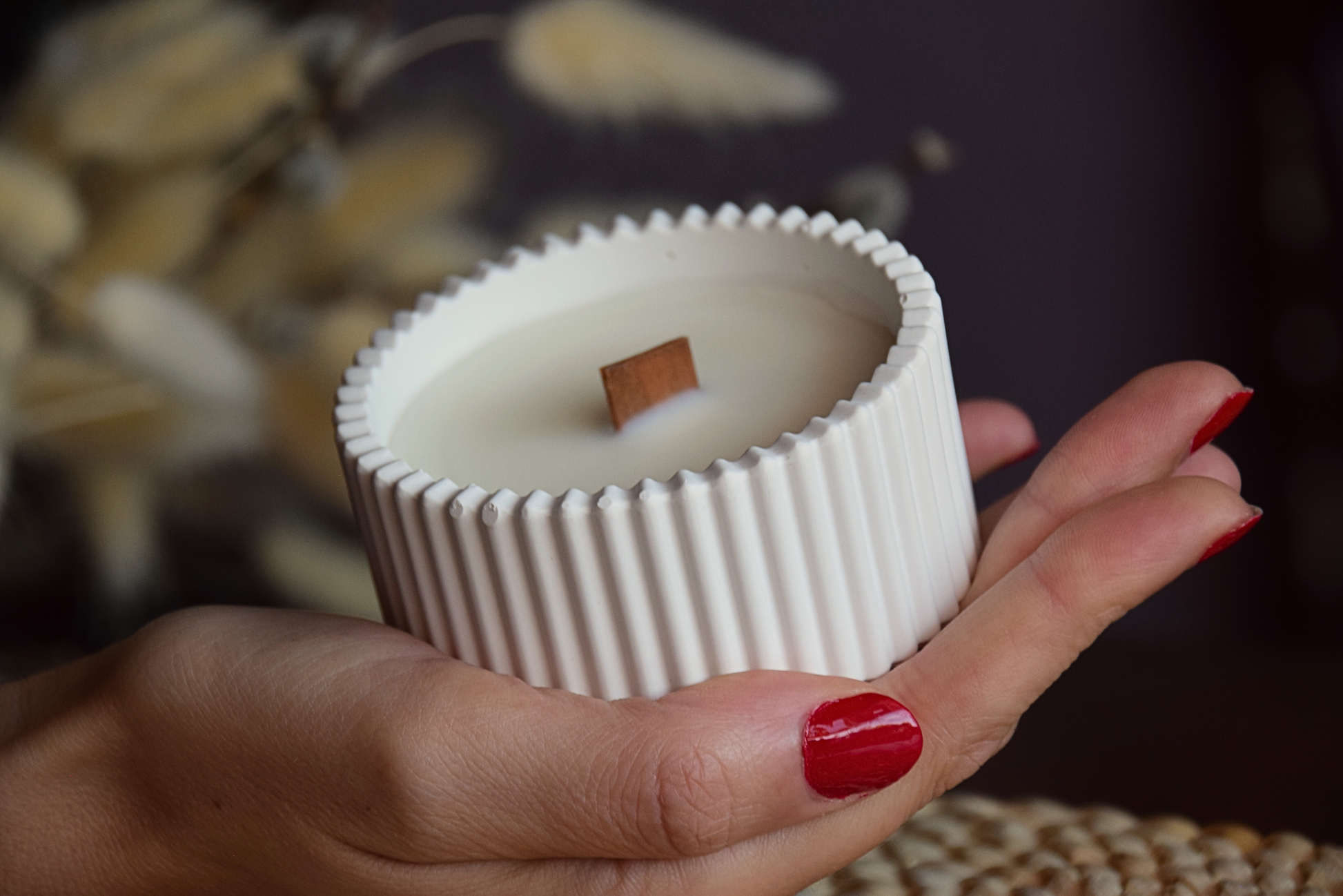 Vela decorativa en cuenco de rallas de tamaño pequeño con mecha de madera sujetado por mano de modelo con fondo rustico. 