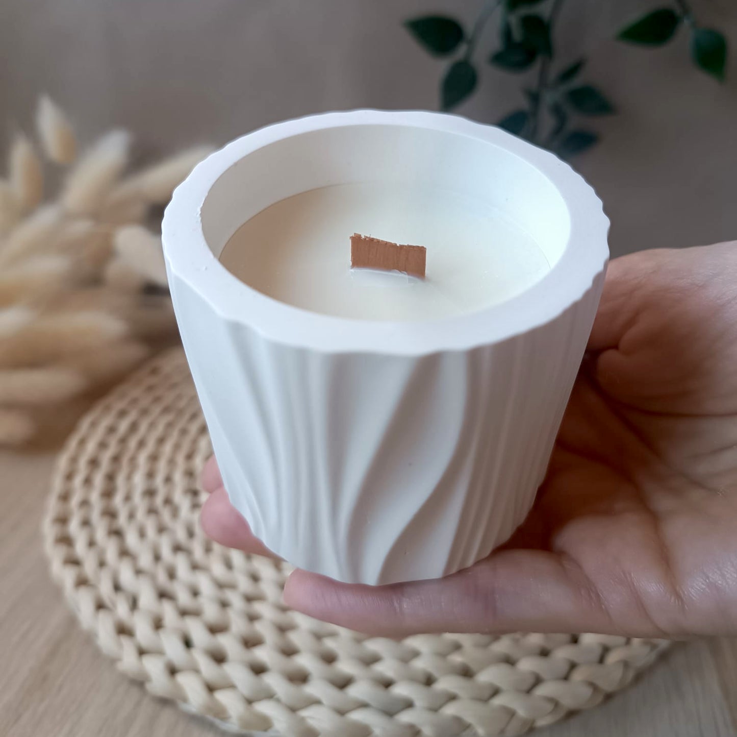 Vela de soja en recipiente de resina mineral blanco con formas orgánicas y mecha de madera sostenida en la mano con fondo neutro