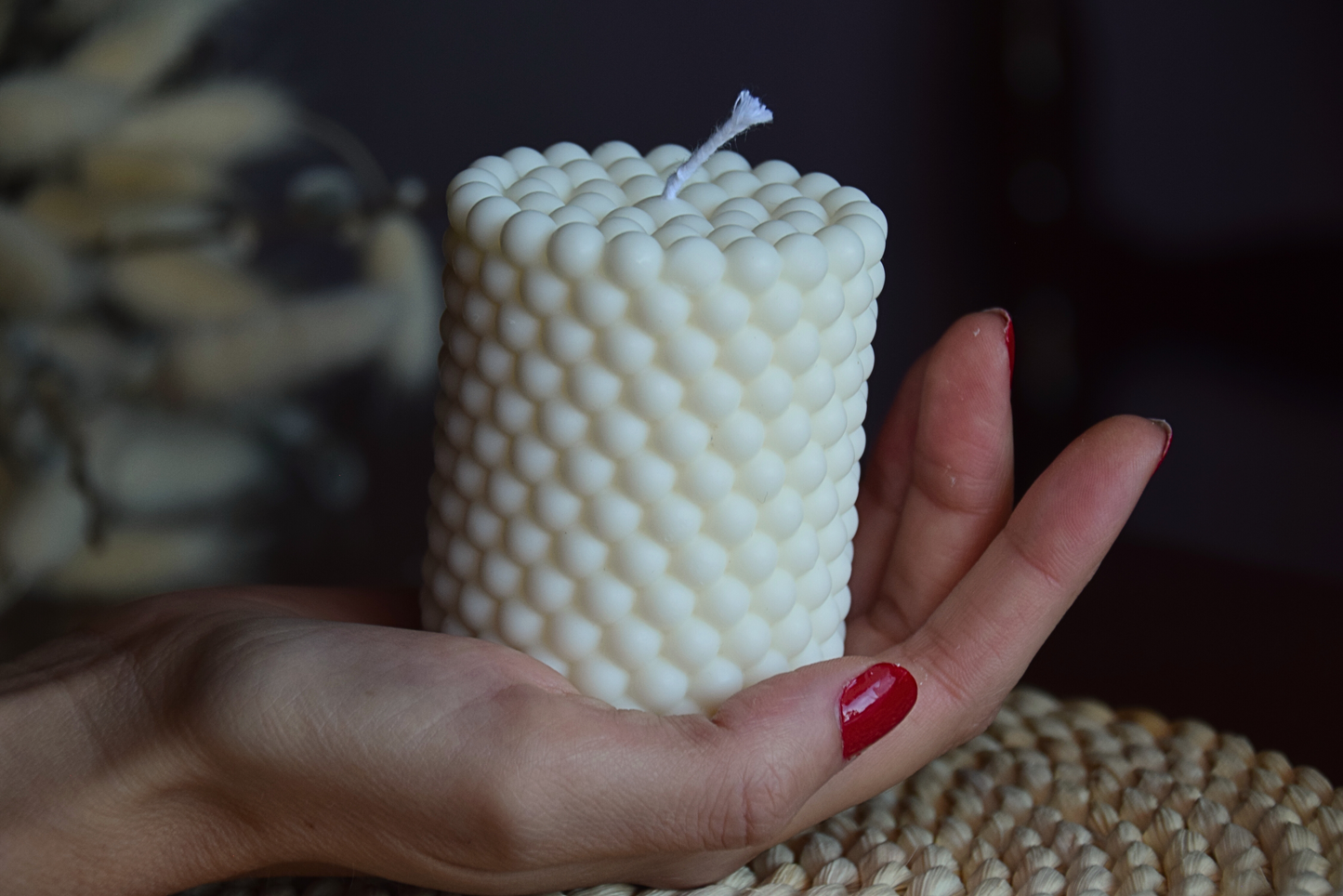Vela decorativa en forma de pilar con burbujas y pabilo de algodon, sujetada por mano de modelo con fondo rustico. 