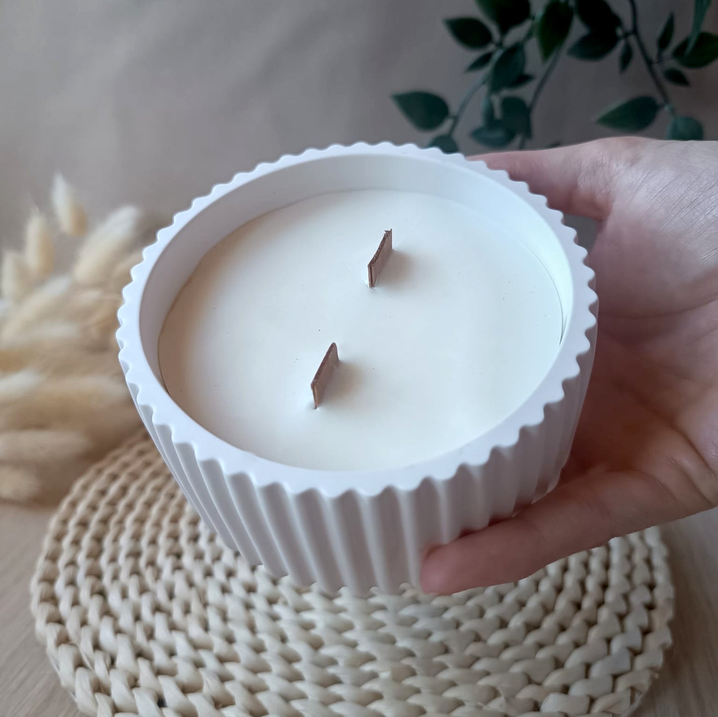 Vela decorativa de soja en bol blanco de resina mineral blanco con textura de rayas con doble mecha de madera sostenida en la mano y fondo neutro