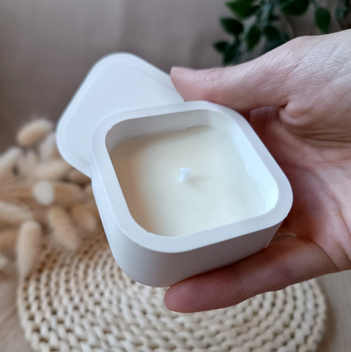 Vela de soja en recipiente cuadrado blanco de resina mineral con tapa y mecha de algodón sostenida en la mano con fondo neutro