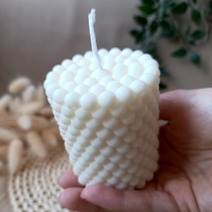 Vela artesanal de soja en forma de cono con relieve de burbujas sostenida en la mano y fondo neutro
