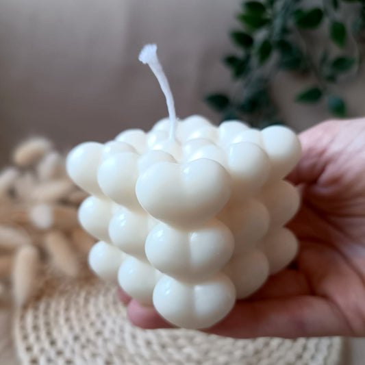 Vela artesanal de soja en forma de cubo con diseño de corazones sostenida en la mano y fondo neutro