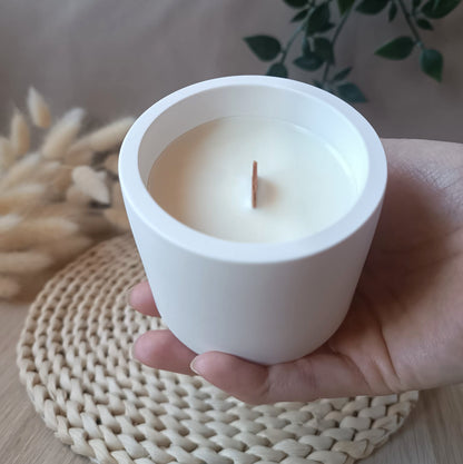 Vela artesanal de soja en recipiente blanco liso de resina mineral con mecha de madera sostenida en la mano y fondo neutro