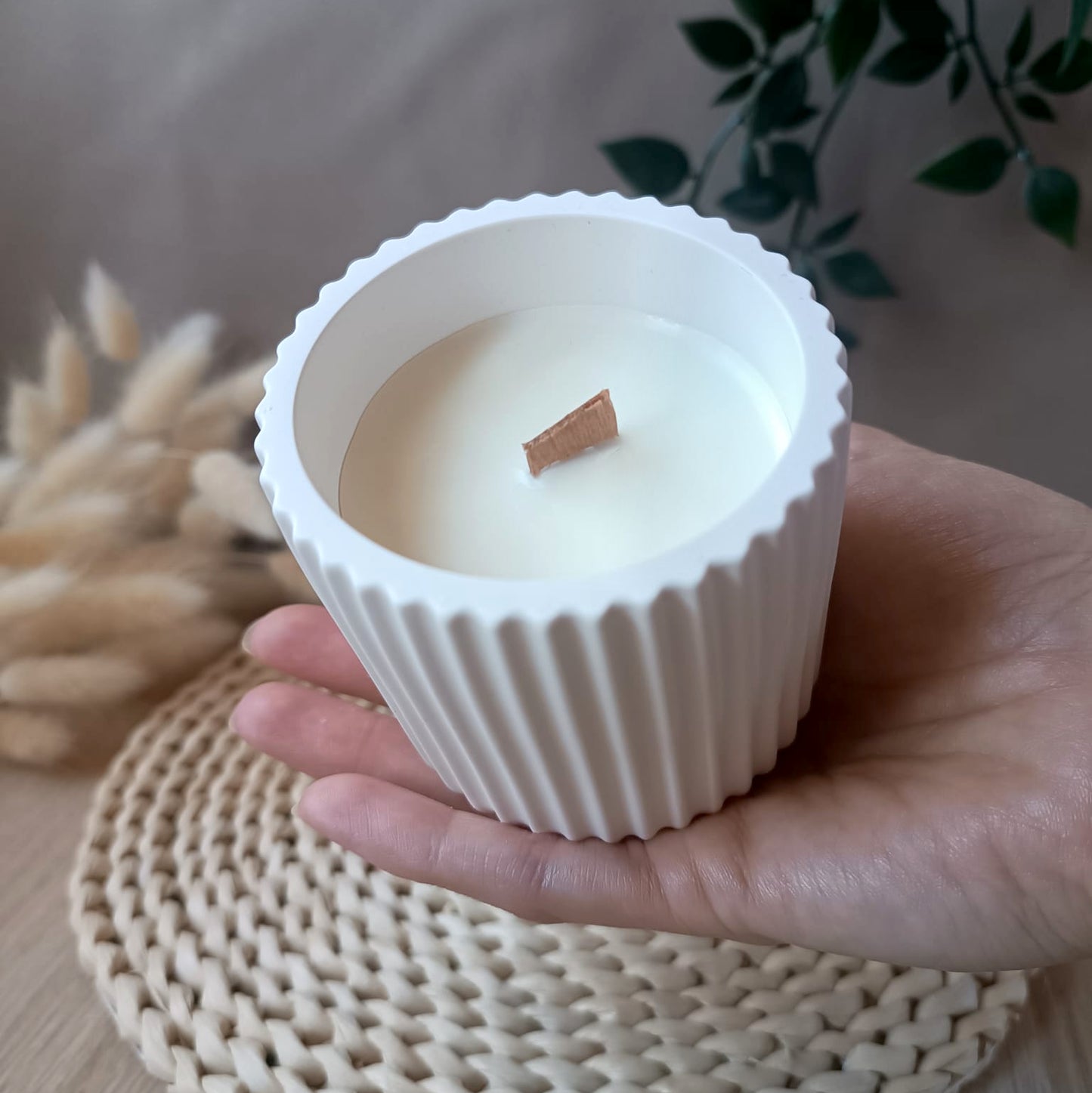Vela artesanal de soja en recipiente blanco de resina mineral con relieve de rayas verticales y mecha de madera sostenida en la mano con fondo neutro