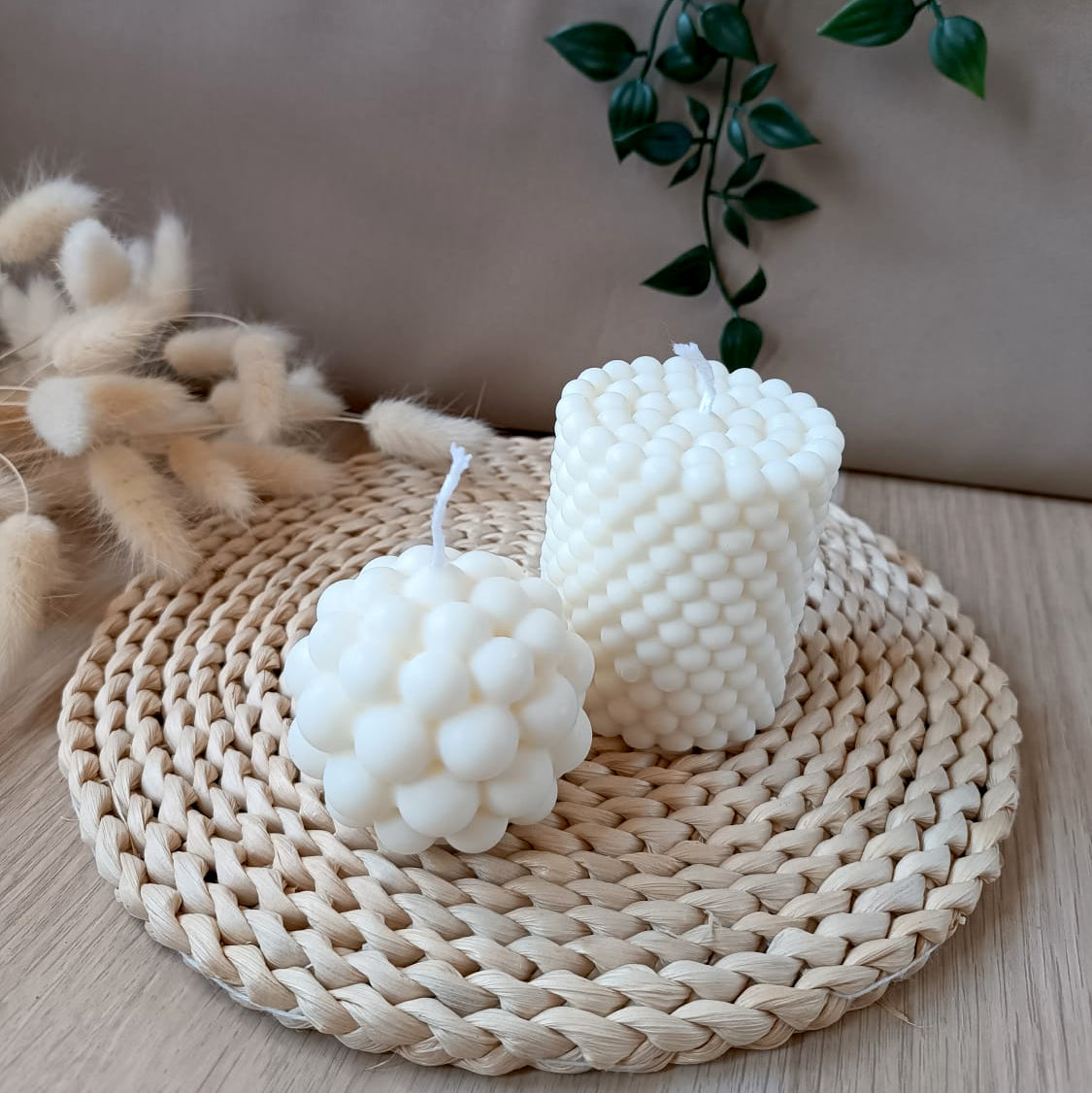 Conjunto de dos velas decorativas de soja con diseño de burbujas una esférica pequeña y otra cilíndrica mas grande sobre base de fibras naturales y fondo neutro