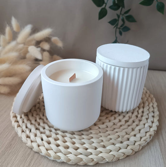 Conjunto de dos velas decorativas de soja presentadas en recipiente de resina mineral uno liso y otro acanalado con tapa y mecha de madera sobre base de fibra natural y fondo neutro