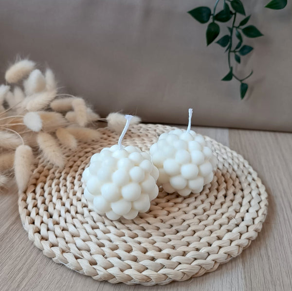 Dos velas decorativas de soja en forma de esfera con diseño de burbujas sobre base de fibras naturales y fondo neutro
