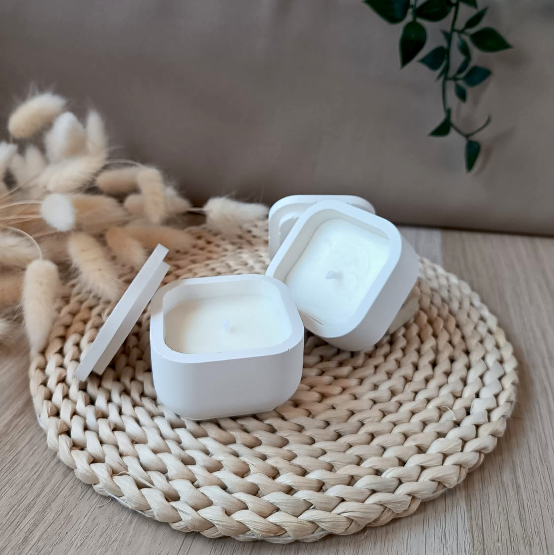 Dos velas de soja en recipiente cuadrado blanco y liso de resina mineral con tapa y mecha de algodón sobre base fibras naturales y fondo neutro