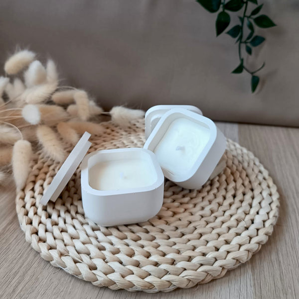 Dos velas de soja en recipiente cuadrado blanco y liso de resina mineral con tapa y mecha de algodón sobre base fibras naturales y fondo neutro