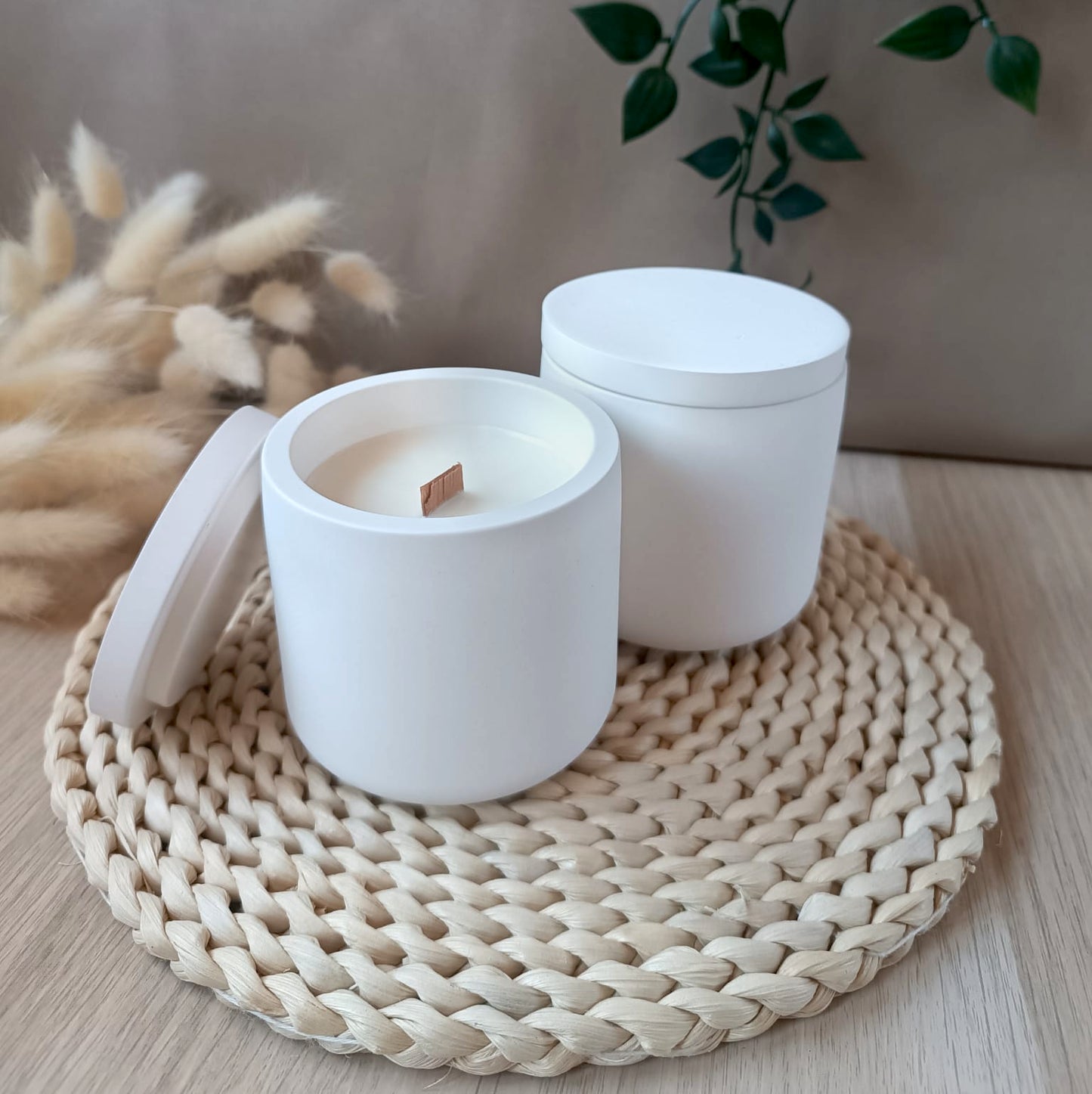 Dos velas de soja en recipiente liso de resina mineral con tapa a juego y mecha de madera sobre base de fibras naturales y fondo neutro
