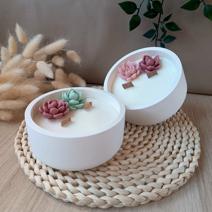Dos velas decorativas de soja en recipiente de resina mineral con doble mecha de madera y suculentas sobre base natural y fondo neutro